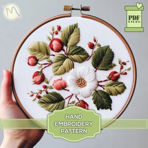 Op de afbeelding: Een handborduurpatroon met een witte bloem met groene bladeren en rode bessen. Het patroon is in een ring en klaar om te worden geborduurd. De tekst "HAND EMBROIDERY PATTERN" staat onderaan de afbeelding.