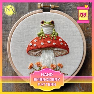 Peut inclure: Modèle de broderie à la main représentant une grenouille verte assise sur un champignon rouge et blanc à pois blancs. Le champignon est entouré de fleurs orange. Le motif est présenté dans un cerceau de broderie en bois.