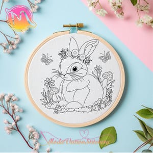 May include: A black and white line drawing of a rabbit wearing a flower crown and sitting in a field of flowers and mushrooms. The image is in a round embroidery hoop.
