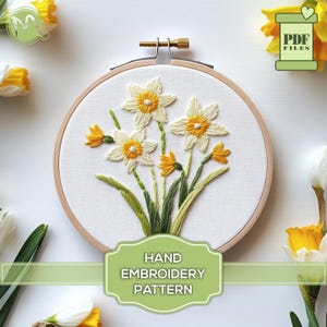 May include: A hand embroidery pattern featuring a bouquet of white daffodils with yellow centres and green leaves. The pattern is displayed in a wooden embroidery hoop. The text "HAND EMBROIDERY PATTERN" is displayed below the hoop.