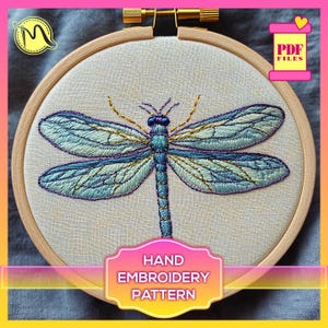 Puede incluir: Un patrón de bordado a mano que presenta una libélula detallada con hilo azul, verde y dorado sobre una tela de lino blanca en un aro de bordado de madera. El texto "HAND EMBROIDERY PATTERN" está impreso en una etiqueta debajo del aro.