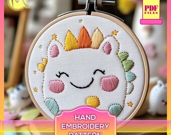 May include: A white embroidery hoop with a hand-stitched design of a smiling cartoon creature wearing a crown. The creature has a yellow, orange, and white crown, green and blue leaves around its head, and pink cheeks. The hoop is on a wooden surface. "HAND EMBROIDERY PATTERN" is written on a label below the hoop.