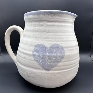 Può includere: Brocca in ceramica bianca con un motivo a forma di cuore blu. La brocca ha un'ampia apertura e un manico.
