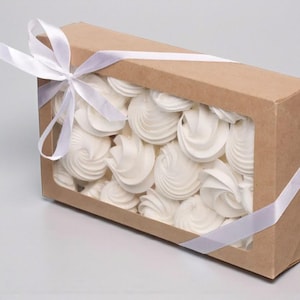 Puede incluir: Una caja rectangular de cartón con una ventana transparente llena de merengues blancos. La caja está atada con una cinta de raso blanca. Las galletas tienen un diseño en espiral.
