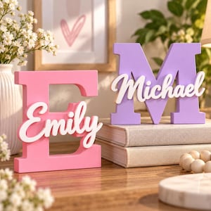 May include: A pink wooden letter E with the white script name Emily attached to the front and a purple wooden letter M with the white script name Michael attached to the front, both standing upright as decorative freestanding block letters.