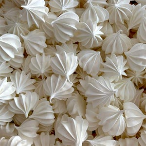 May include: A close-up of a collection of white meringue cookies. Each cookie has a unique shape, with a fluted, fan-like design. The meringues are a brilliant white, and the image is well-lit, highlighting the texture and details of each cookie.