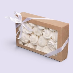 Peut inclure: Une boîte rectangulaire en carton avec une fenêtre transparente remplie de meringues blanches tourbillonnantes. La boîte est attachée avec un ruban de satin blanc en forme de nœud. Les meringues sont un dessert léger et aéré.