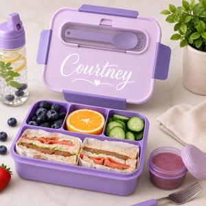 May include: A rectangular lavender bento box with a matching lid featuring the name Courtney in white script. The container has three internal compartments holding blueberries, orange slices, and cucumber rounds, plus two sandwiches.