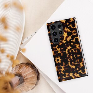 Puede incluir: Una funda de tel&eacute;fono de color marr&oacute;n y negro de caparaz&oacute;n de tortuga con un recorte para el objetivo de la c&aacute;mara. La funda tiene un patr&oacute;n texturizado.