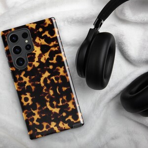 Puede incluir: Una funda de tel&eacute;fono negra con un patr&oacute;n de caparaz&oacute;n de tortuga. La funda est&aacute; sobre una superficie blanca junto a unos auriculares negros.