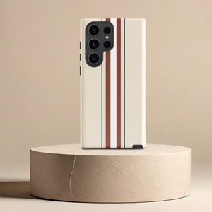 May include: A cream-colored phone case with three vertical stripes in burgundy, white, and navy blue. The case features a multi-lens camera system and is displayed on a beige cylindrical pedestal.