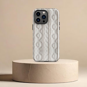 May include: A white phone case with a cable knit pattern. The case has a black camera lens with three lenses.