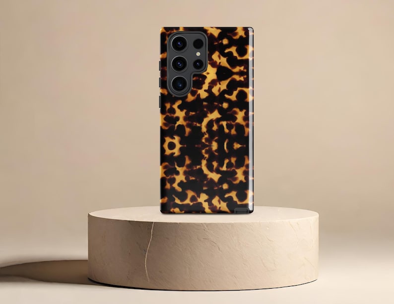 Puede incluir: Una funda de tel&eacute;fono negra con un patr&oacute;n de caparaz&oacute;n de tortuga. La funda est&aacute; sobre un pedestal de color marr&oacute;n claro.