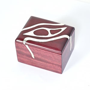 May include: A small wooden box with a dark red finish and a white inlay design. The design features a stylized eye shape with wavy lines.