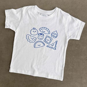 May include: A white short-sleeved t-shirt featuring a blue graphic print of various dumplings, each with a simple smiling face. The design includes different shapes of filled dough, such as round, triangular, and pleated varieties, arranged centrally.