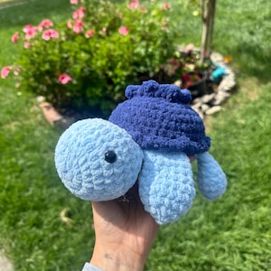 May include: A handmade crochet turtle in shades of blue. The turtle has a light blue head and limbs, a dark blue shell, and a black eye. The plush toy is held in a hand against a background of green grass and pink flowers.