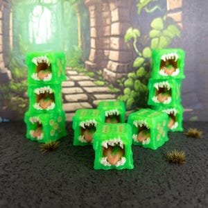 May include: Green, cube-shaped dice with open mouths, white teeth, and pink tongues. The dice are stacked in groups and scattered on a dark surface. The dice have a cartoonish, monster-like appearance.