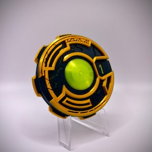 May include: A circular, futuristic-looking device with a black and gold design. The device has a bright green center and the number "1" is visible. It is displayed on a clear plastic stand.