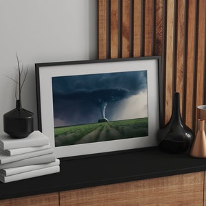 May include: A framed photograph of a tornado in a field. The photo is in a black frame and is sitting on a black shelf with a stack of white books, a black vase, and a gold and gray vase.