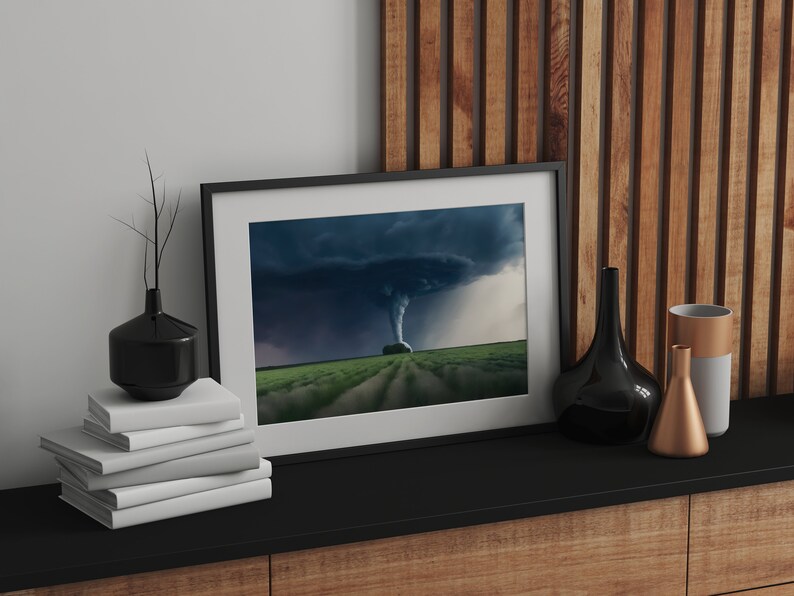 May include: A framed photograph of a tornado in a field. The photo is in a black frame and is sitting on a black shelf with a stack of white books, a black vase, and a gold and gray vase.