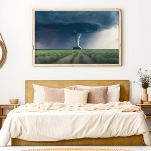 May include: A bedroom interior with a gold headboard bed, white bedding, and a framed photograph of a tornado over a field of green grass. The room is decorated with a round woven mirror, a woven basket, and a wooden side table with a vase of dried flowers.