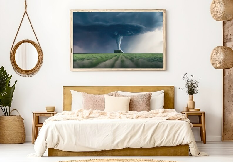 May include: A bedroom interior with a gold headboard bed, white bedding, and a framed photograph of a tornado over a field of green grass. The room is decorated with a round woven mirror, a woven basket, and a wooden side table with a vase of dried flowers.