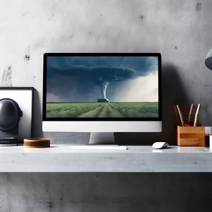 May include: A modern home office workspace with a desktop computer displaying a dramatic image of a tornado over a green field. The desk is made of light gray concrete and has a black speaker, a wooden bowl, and a lamp with a black shade.