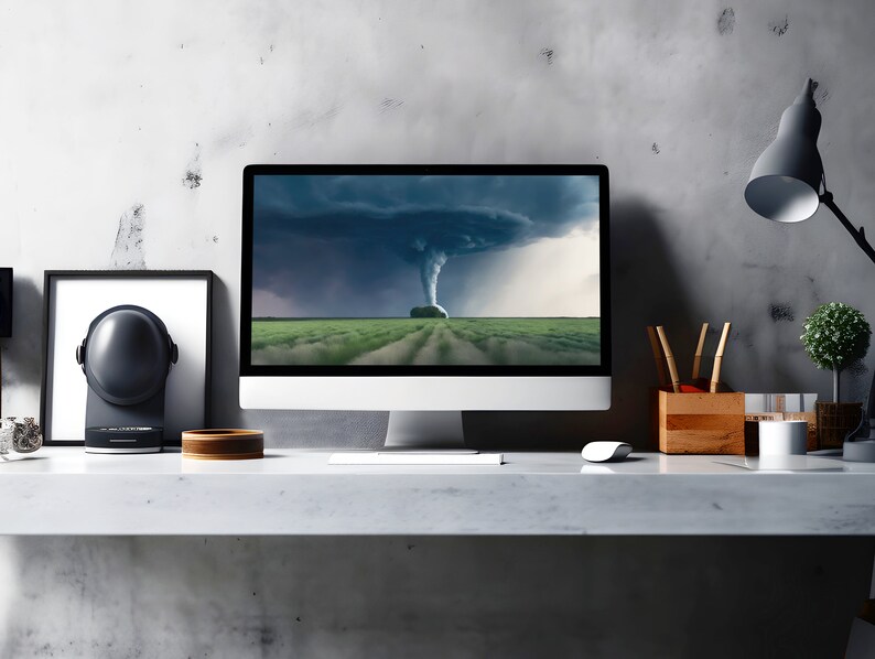 May include: A modern home office workspace with a desktop computer displaying a dramatic image of a tornado over a green field. The desk is made of light gray concrete and has a black speaker, a wooden bowl, and a lamp with a black shade.