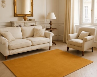 May include: A rectangular, mustard-colored rug dominates the foreground of a living room scene. The rug is placed on a herringbone-patterned wood floor. A cream-colored sofa and armchair are visible, along with a gold-framed mirror and a table lamp.