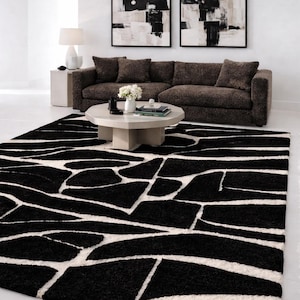 May include: A contemporary living room with a striking black and white patterned rug. The rug features a bold geometric design. A brown sofa with cushions is placed against a white wall, with abstract art and a side table with a lamp.