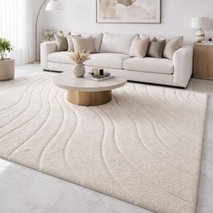 May include: A cream-coloured, rectangular area rug with a wave pattern. The rug is placed on a marble floor in a living room setting. A white sofa with beige cushions, a round coffee table, and framed artwork are also visible.