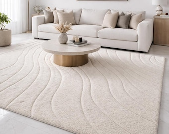 May include: A cream-colored, rectangular area rug with a wave pattern. The rug is placed on a marble floor in a living room setting. A white sofa with beige pillows, a round coffee table, and a framed artwork are also visible.