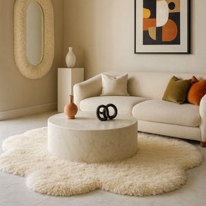 Op de afbeelding: Een moderne woonkamer met een crèmekleurige bank, een ronde salontafel en een zacht, crèmekleurig vloerkleed. Een decoratieve spiegel en abstracte kunst sieren de muren. Accentkussens in olijf en oranje voegen kleur toe.