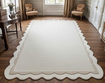 May include: A rectangular, off-white rug with a scalloped edge. The rug has a textured surface and a border in a lighter beige color. The rug is placed on a dark wood floor in a room with neutral-toned walls and furniture.
