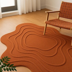 May include: An orange rug with a distinctive, organic form and a topographic pattern. The rug has raised, curved lines that create a layered effect. A wooden chair with a woven seat is nearby, complementing the rug's warm tones. The rug is a decorative floor covering.