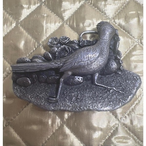 May include: A silver-colored metal belt buckle featuring a detailed roadrunner bird standing on a rocky surface with foliage. The buckle has a classic western design, with a sturdy metal loop for the belt.