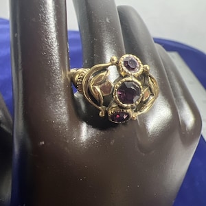 May include: A gold-toned ring with three large, round, deep purple gemstones and smaller accent stones. The ring features an ornate design with leaf and floral details. The ring is displayed on a dark brown hand-shaped display.