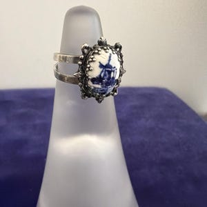 May include: A silver ring featuring an oval ceramic inlay with a blue and white windmill design. The setting has a decorative beaded edge and a split band design. The ring is displayed on a translucent cone-shaped stand for clear visibility.