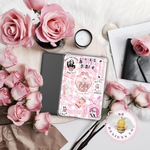 May include: A gray Amazon Kindle next to a pink and white patterned case with stickers. The stickers include a skeleton, ghost, and text that reads "emotional support kindle" and "spooky bookclub". Pink roses and a small candle are also in the image.