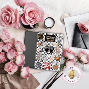 May include: A gray Amazon Kindle beside a clear case decorated with stickers. The case has a black and white checkered pattern and the text "Emotional Support Kindle". Pink roses and a candle surround the Kindle.