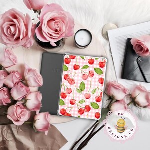May include: An Amazon Kindle e-reader with a cherry-patterned cover rests beside a gray Kindle and pink roses. The cover features red cherries and green leaves on a pink and white gingham background. A small candle and a black wallet are also present.