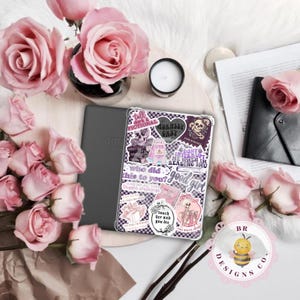 May include: A gray Amazon Kindle e-reader with a clear case covered in stickers, including text that reads "tall dark fictional" and "morally grey." Pink roses and a small candle surround the e-reader.