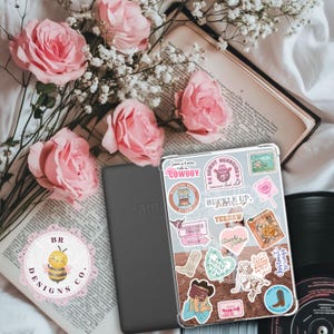 May include: An Amazon Kindle e-reader with a clear case covered in cowboy-themed stickers, resting on an open book. Pink roses and white baby's breath flowers are arranged nearby. A vinyl record is in the background. The logo "BR DESIGNS CO." is visible.