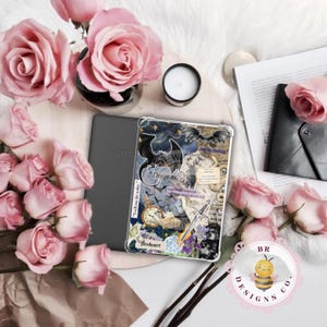 May include: A clear Kindle case with a collage design featuring dragons, stars, and text. The case is next to a gray Kindle device, pink roses, a candle, and other decorative items. The text on the case includes "Dragons" and "fireheart."