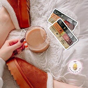 May include: A relaxed image showing a clear glass mug with a light brown drink, and book stickers. The stickers display book titles and author names. The person is wearing brown slippers with white faux fur trim.