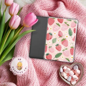 May include: A pink Kindle case with a strawberry pattern, resting on a pink knitted blanket. The case features a repeating design of strawberries, leaves, and small white flowers. A heart-shaped bowl of marshmallows and pink tulips are also in the image.