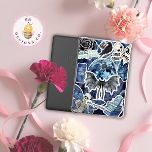 May include: A black e-reader with a decorative case featuring a blue and white design with a bat, moon, and skull. The case has text that reads "shh, I'm reading", "Smut again?", and "hello, fayre darling". Pink flowers and ribbons are in the background.