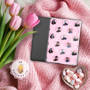 Puede incluir: Una funda rosa para Kindle con una cubierta transparente con imágenes de dibujos animados de personajes de películas de terror. La funda está sobre una manta de punto rosa, junto a un Kindle, tulipanes rosas y un cuenco en forma de corazón con malvaviscos.