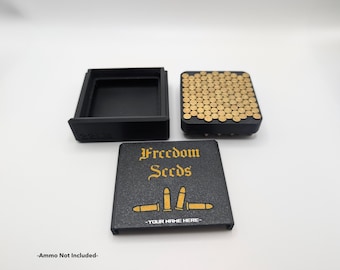 Könnte beinhalten: Schwarze Box mit einer goldenen Aufschrift "Freedom Seeds" und einer Grafik von drei goldenen Kugeln. Die Box ist geöffnet und der Deckel ist separat. Die Box ist mit goldfarbenen Pellets gefüllt.