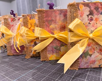 May include: Four small, decorative books with floral designs and burlap covers. Each book is tied with a yellow satin ribbon bow.